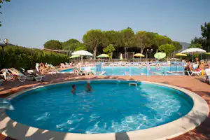 Camping-Pool mit blauem Wasser, Liegen und Sonnenschirmen, dahinter Zaun und hohe Bäume im Hintergrund.
