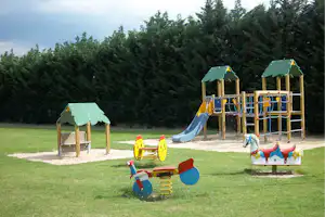 Spielplatz mit Kletterturm und Rutsche, Sandfläche und Spielgeräten auf dem Campinggelände, umgeben von Bäumen.