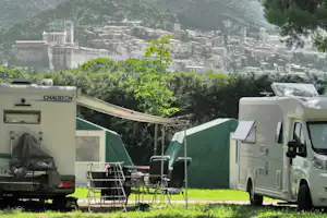 Zwei Wohnmobile auf einem Campingplatz mit grünen Zelten, Campingstühlen und Blick auf ein Bergdorf im Tal.