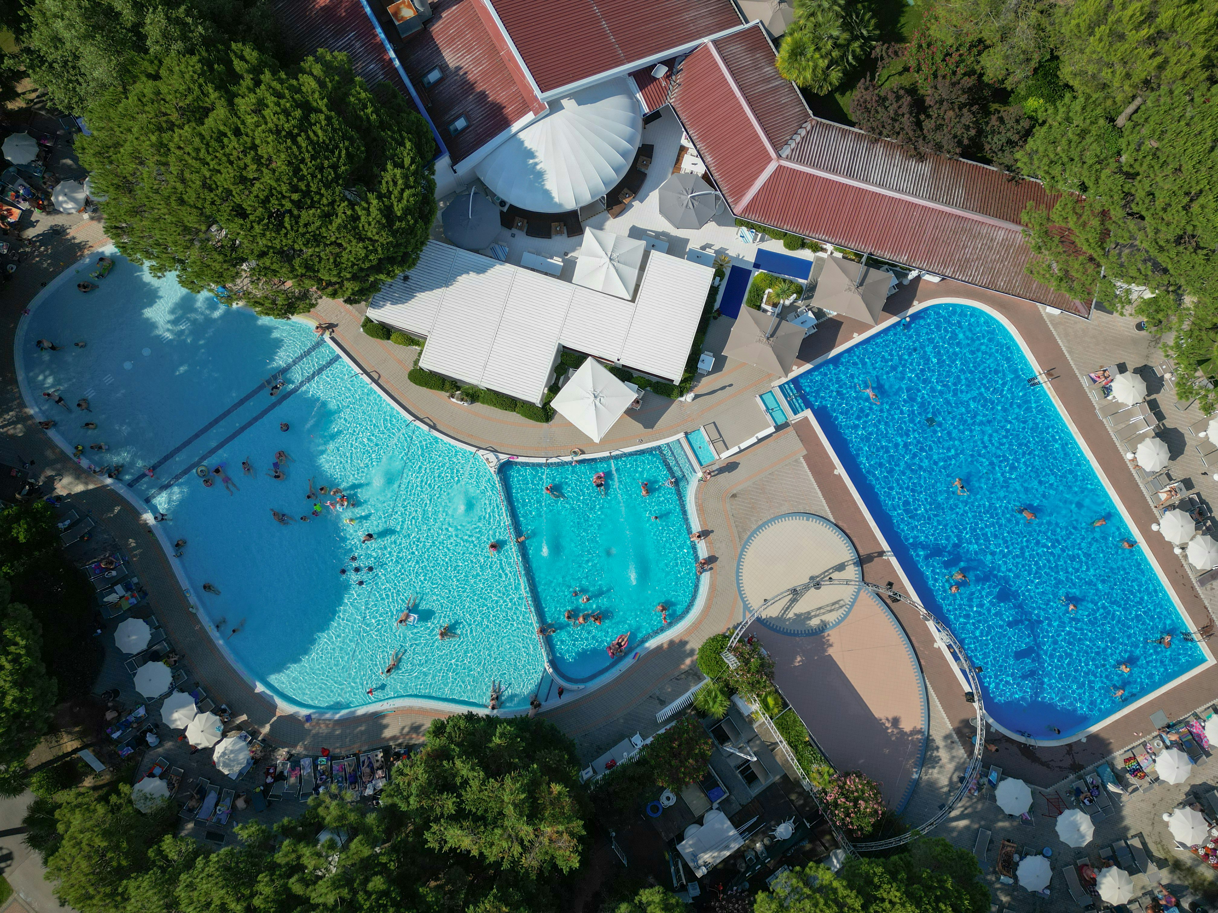 Luftaufnahme von zwei großen Camping-Pools mit Liegen, Sonnenschirmen und Wegen, umgeben von Bäumen und Gebäuden.