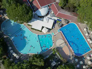 Luftaufnahme von zwei großen Camping-Pools mit Liegen, Sonnenschirmen und Wegen, umgeben von Bäumen und Gebäuden.
