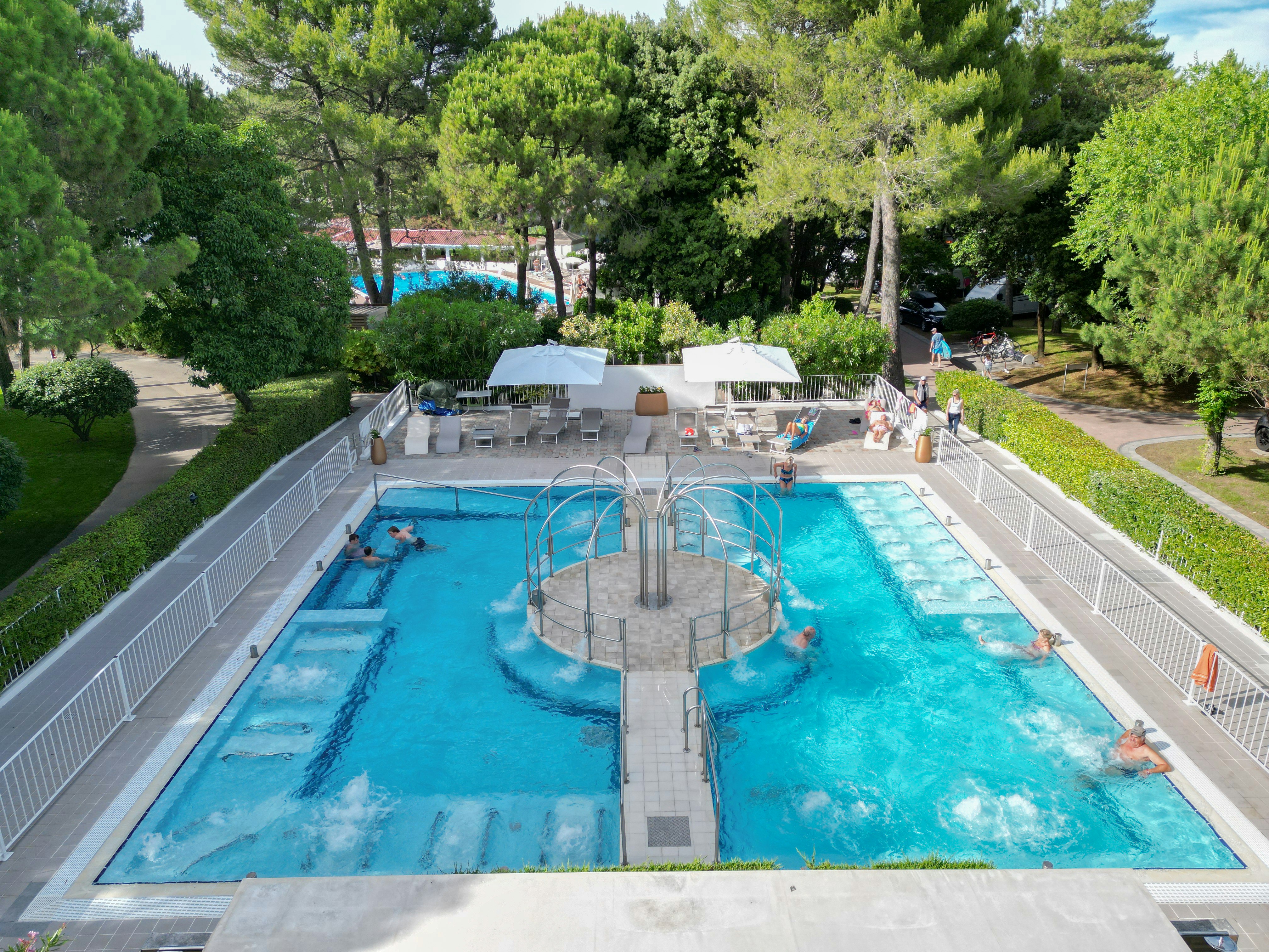Großer Camping-Pool mit Rundbecken, Liegen und Sonnenschirmen, umgeben von Hecken und Bäumen.