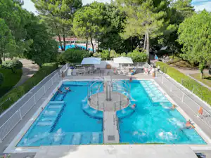 Großer Camping-Pool mit Rundbecken, Liegen und Sonnenschirmen, umgeben von Hecken und Bäumen.