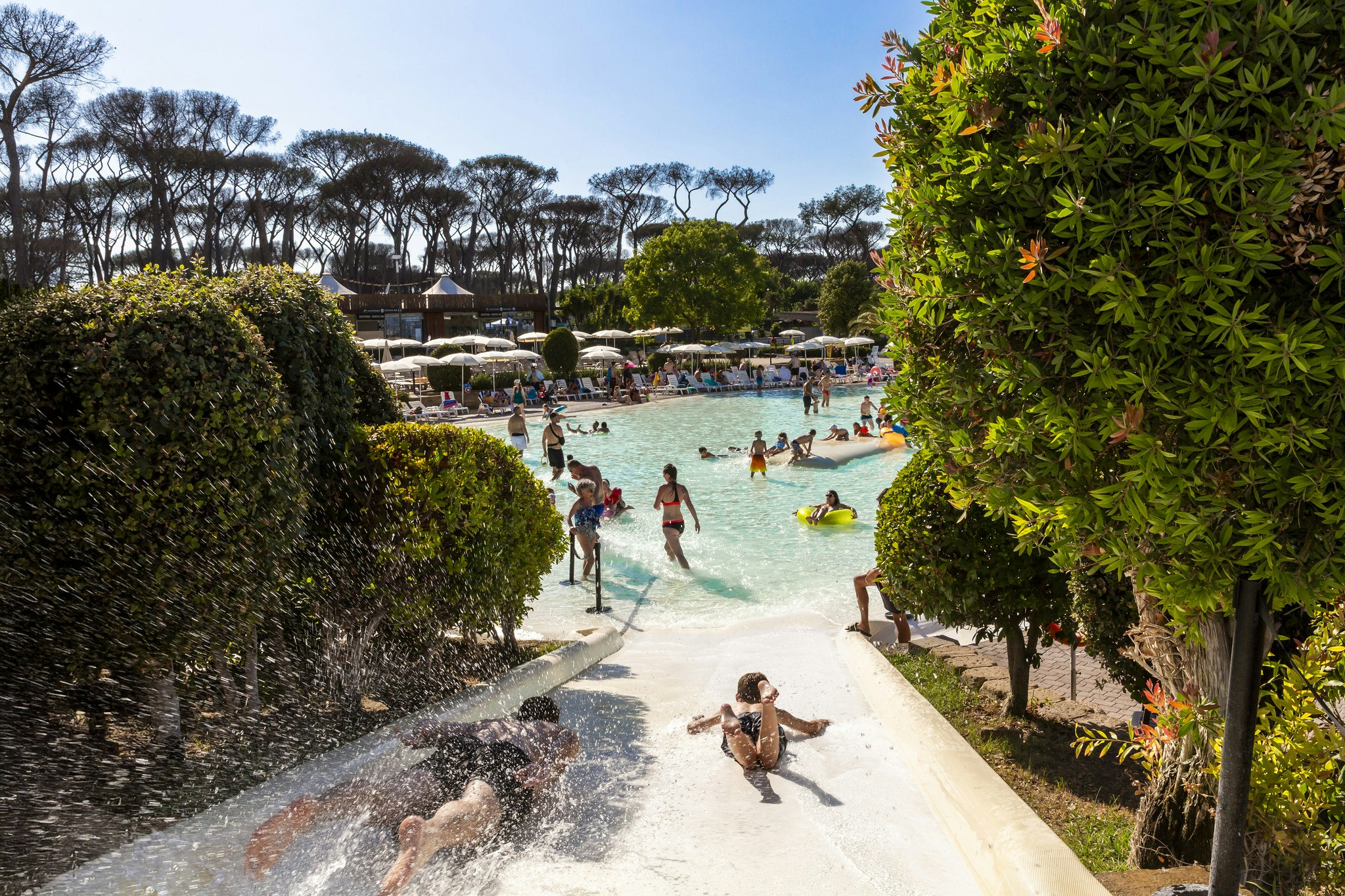 Schwimmbad mit türkisfarbenem Wasser, Rutschen und mehreren Personen; im Hintergrund Bäume und gepflegte Wege zwischen Hecken.
