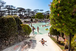 Schwimmbad mit türkisfarbenem Wasser, Rutschen und mehreren Personen; im Hintergrund Bäume und gepflegte Wege zwischen Hecken.