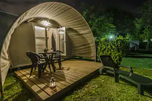 Beiges Zelt auf einer Holzterrasse mit Tisch und Stühlen, daneben Gartenliegen und Büsche im abendlichen Campingbereich.