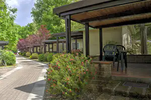Bungalow mit überdachter Terrasse, Gartenblumen und Sitzgarnitur, daneben ein gepflasterter Weg zwischen weiteren Gebäuden.