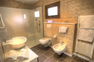 Helles Badezimmer mit Dusche, Waschbecken, zwei Toiletten und Handtüchern, Fliesenboden und Fenster im Hintergrund.