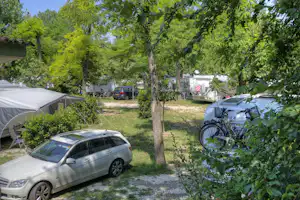 Campingplatz mit geparktem Kombi, Zelt und Wohnwagen zwischen Bäumen, Kiesweg und Rasenfläche.