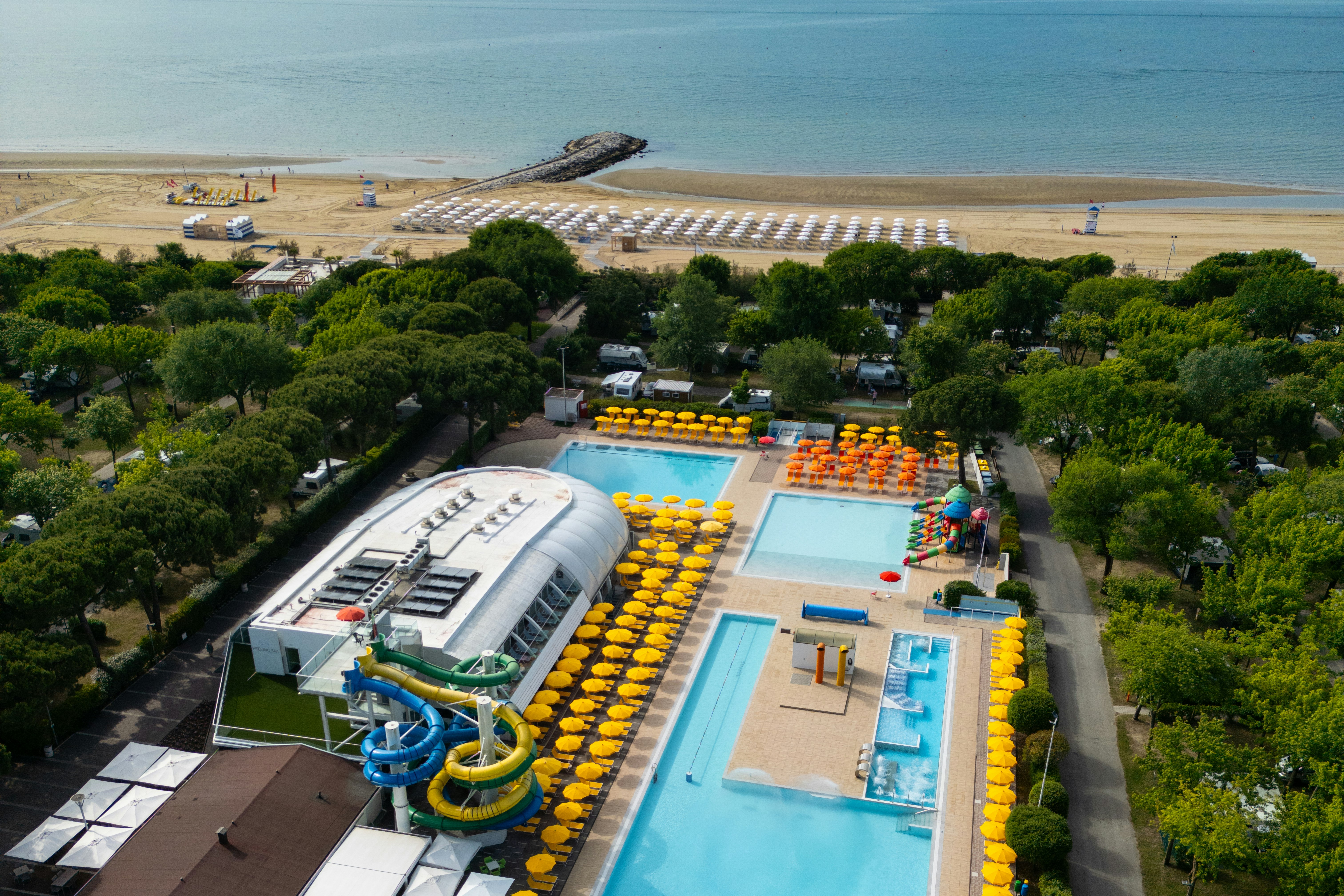 Großes Campinggelände mit zwei Pools, vielen gelben und orangefarbenen Liegen, Bäumen und Blick auf den Strand und das Meer.