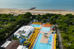 Großes Campinggelände mit zwei Pools, vielen gelben und orangefarbenen Liegen, Bäumen und Blick auf den Strand und das Meer.