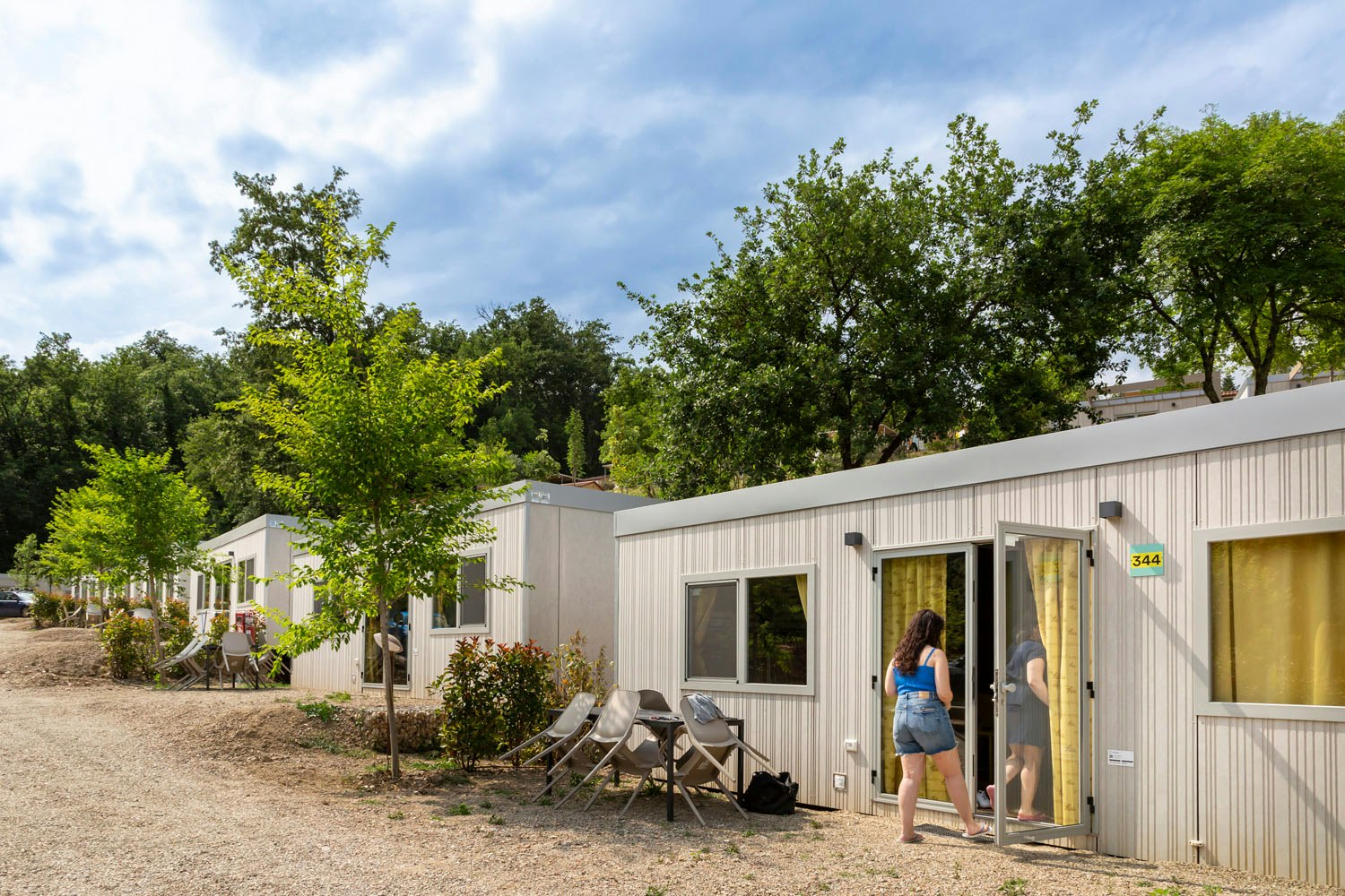Camping-Bungalow mit Nummer 344, Terrasse mit Stühlen und Tisch, umgeben von Bäumen und Kiesweg.
