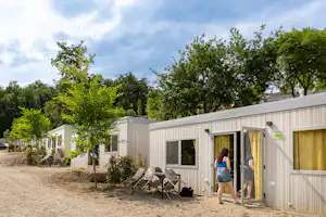 Camping-Bungalow mit Nummer 344, Terrasse mit Stühlen und Tisch, umgeben von Bäumen und Kiesweg.