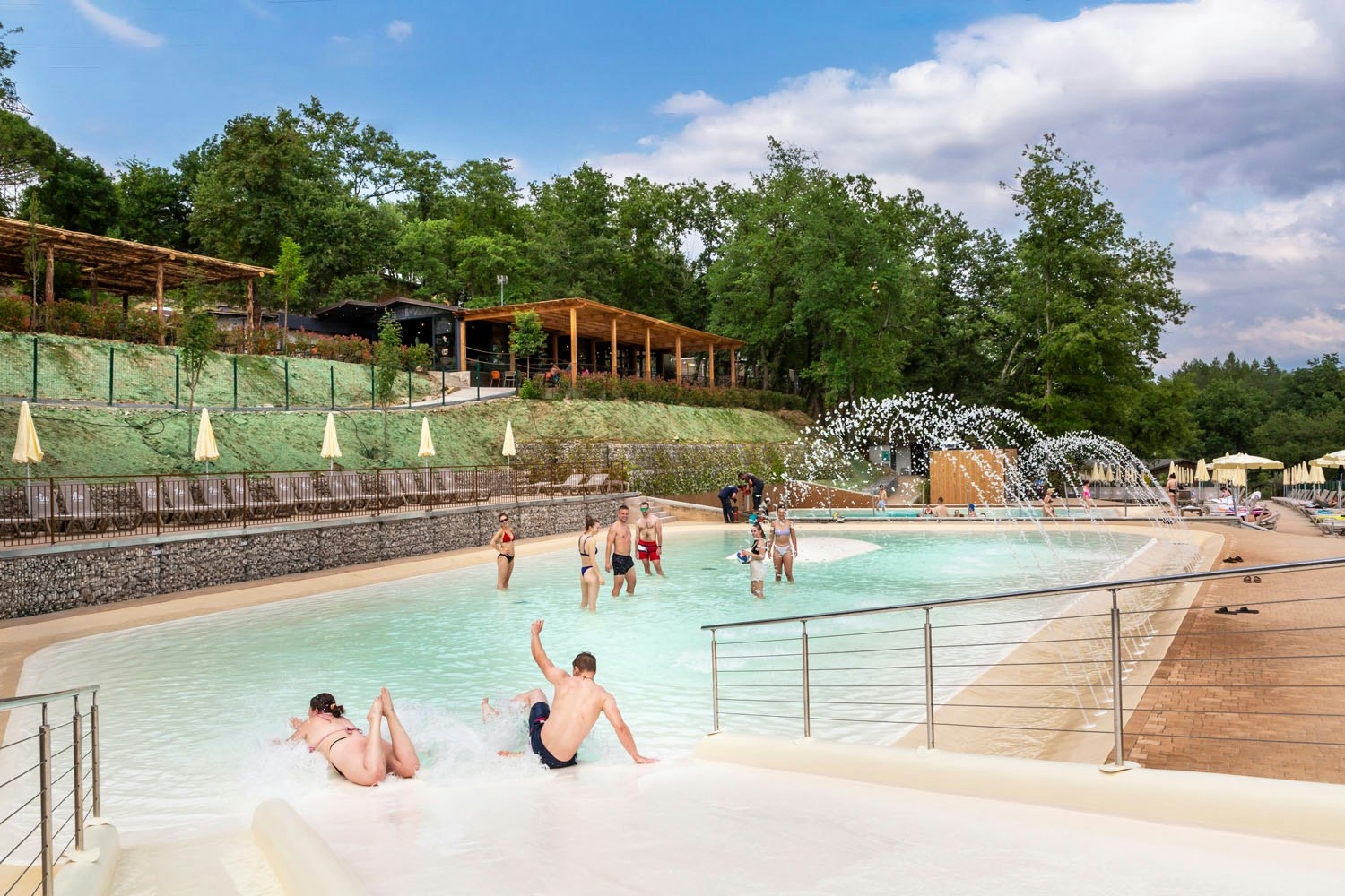 Beheizter Camping-Pool mit mehreren Personen im Wasser, Sonnenschirmen am Beckenrand und Holzterrasse im Hintergrund.
