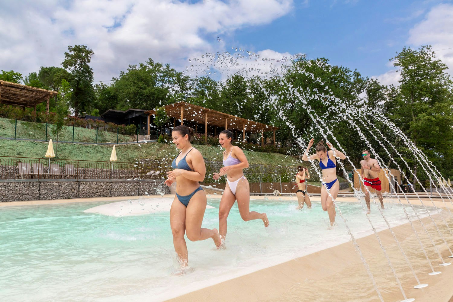 Schwimmbecken im Campingbereich mit Wasserfontänen, mehrere Personen in Badebekleidung und Bäume im Hintergrund.