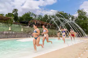 Schwimmbecken im Campingbereich mit Wasserfontänen, mehrere Personen in Badebekleidung und Bäume im Hintergrund.