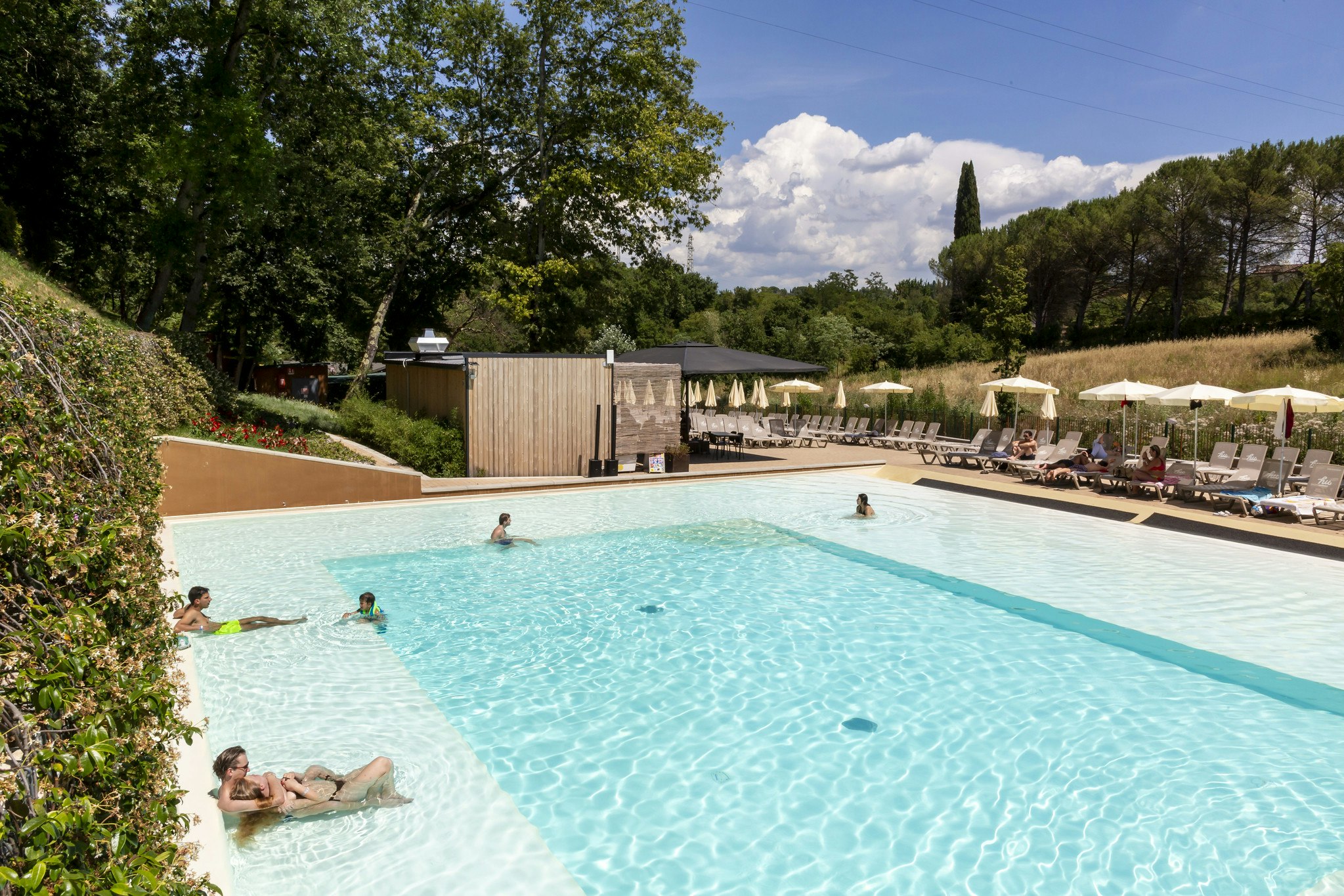 Großes Schwimmbecken mit türkisfarbenem Wasser, Liegen und Sonnenschirmen am Pool, umgeben von Bäumen und Hecken.