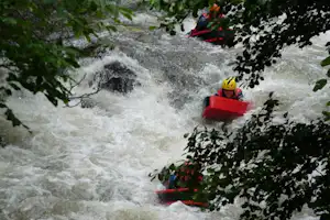 Rote Schlauchboote fahren durch einen Wasserfall, umgeben von grünem Wald und Felsen, mit Wasserrauschen im Vordergrund.