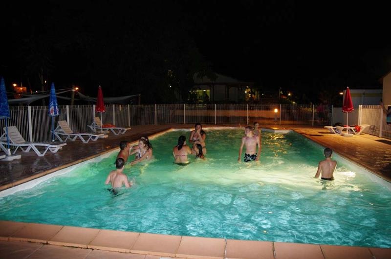 Beleuchteter Camping-Pool in der Nacht mit Kindern im Wasser, Liegestühlen am Rand und Zaun im Hintergrund.