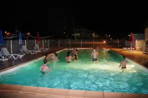 Beleuchteter Camping-Pool in der Nacht mit Kindern im Wasser, Liegestühlen am Rand und Zaun im Hintergrund.