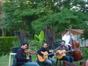 Musikgruppe mit Gitarren und Kontrabass auf dem Campingplatz vor Bäumen und einem kleinen Gebäude im Grünen.
