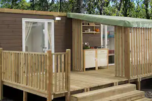 Holz-Campingunterkunft mit Terrasse, überdachtem Eingang und Küchenzeile im Inneren, umgeben von Bäumen.