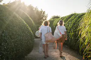 Zwei Personen gehen auf einem Kiesweg zwischen hohen Hecken, mit Rucksäcken und Taschen in der Abendsonne.