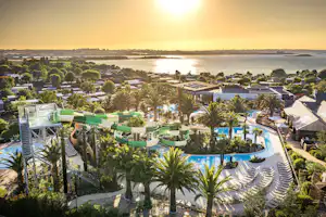 Blick auf einen Campingplatz mit Pool, Liegen und Palmen, im Hintergrund Wasser und eine Küstenlinie bei Sonnenuntergang.