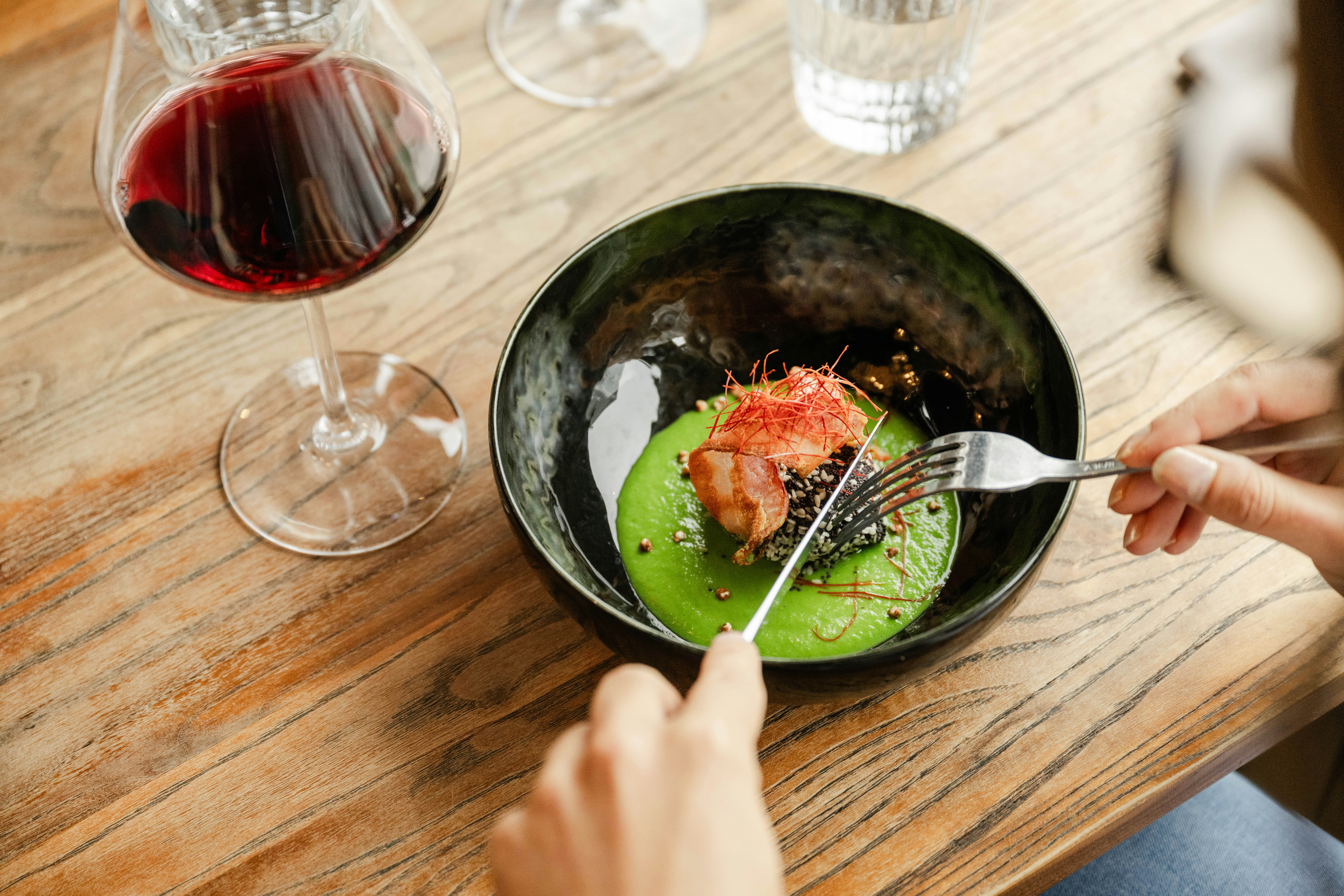 Eine Person isst aus einer Schüssel mit Avocado und Lachs, daneben ein Glas Rotwein auf einem Holztisch.