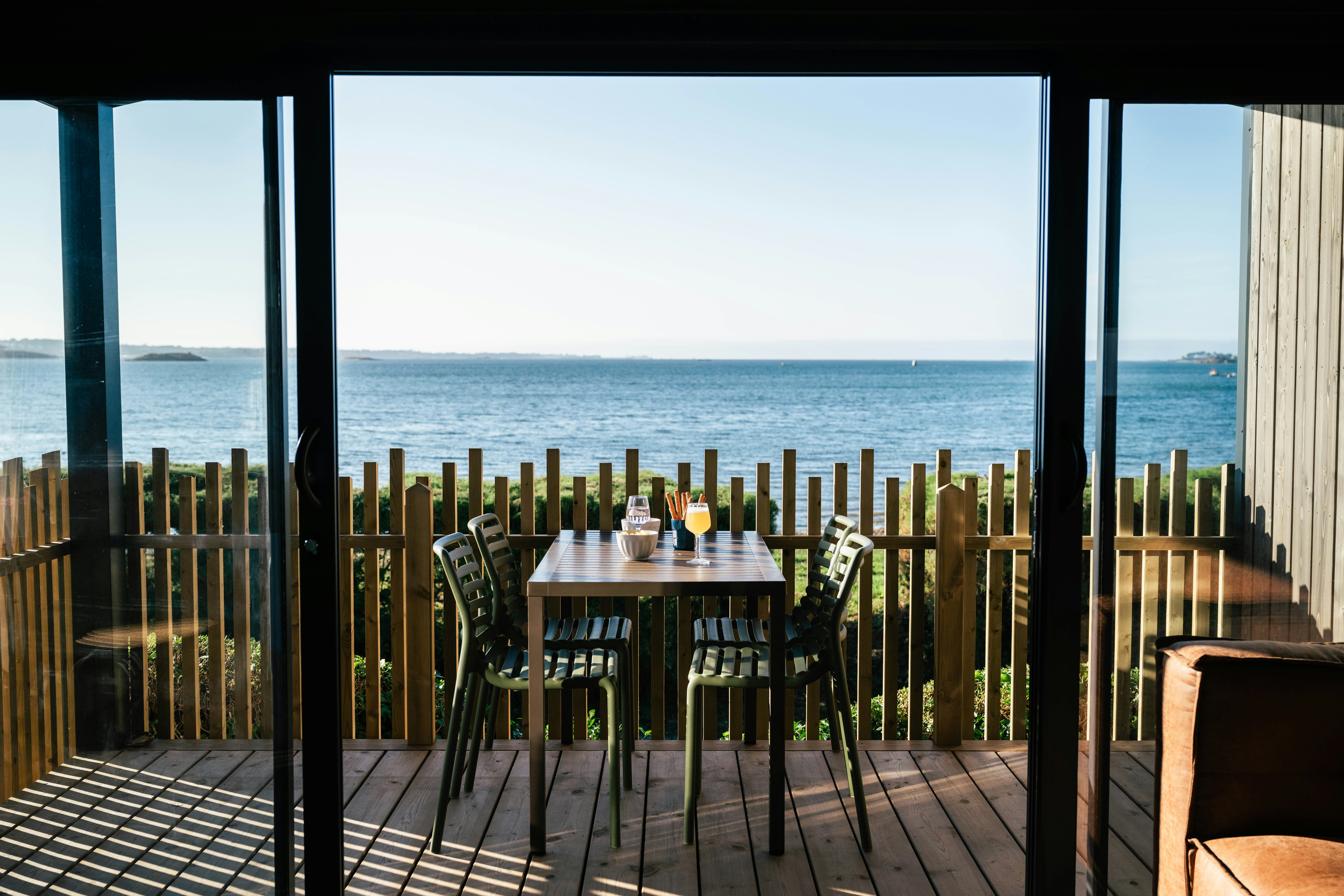 Terrasse mit Holzzaun und Tisch mit Stühlen, Blick auf das Meer durch offene Pergola, am Strand in der Ferne.