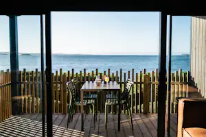 Terrasse mit Holzzaun und Tisch mit Stühlen, Blick auf das Meer durch offene Pergola, am Strand in der Ferne.