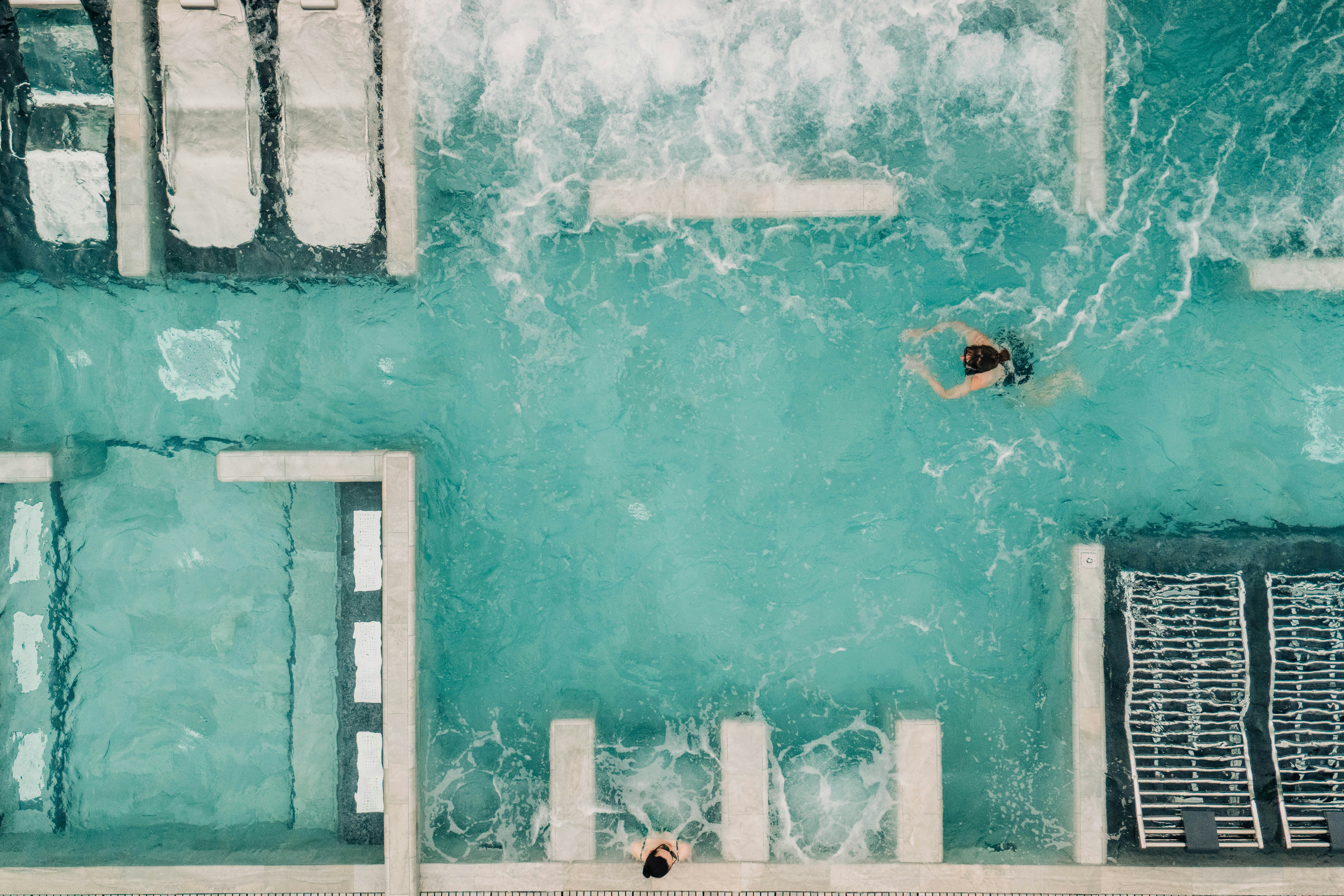 Schwimmbecken mit türkisfarbenem Wasser, Leiter und Sprungbereich; im Hintergrund ein Poolrand und ein weiteres Beckenbecken.