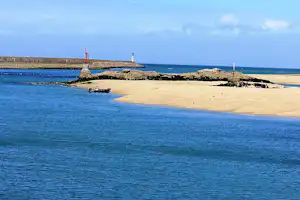 Sandstrand mit Wellenbrechern und Leuchtturm am Meer, im Hintergrund ein weiterer Leuchtturm auf der Mole.