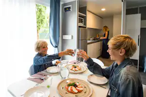 Familie beim Essen im Wohnbereich: Kinder stoßen mit Gläsern an, gedeckter Tisch und Blick in die Küchenzeile.