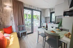 Wohnbereich mit Esstisch, Weingläsern und Küche, große Glastür zum Balkon mit Blick auf Bäume und Sitzplätze.