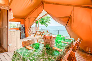Campingzelt mit orangefarbenen Planen, Tisch mit grünem Geschirr und Blick auf das Meer von der Holzterrasse.