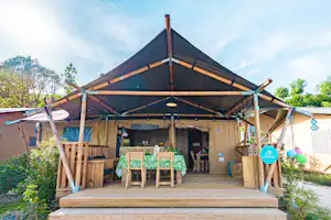 Glamping-Zelt mit Holzterrasse, Tisch und Sitzbank, Holzpfosten und Markise unter blauem Himmel.