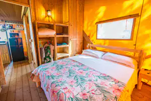 Gemütliches Schlafzimmer im Holzbungalow mit Doppelbett, farbiger Bettwäsche, Schrank und Fenster mit Vorhang.