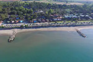 Luftaufnahme eines Campingplatzes am Strand mit Steinschutzdamm, Uferpromenade und mehreren Gebäuden im Grünen.