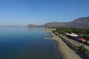 Blick auf den Strand am Meer mit Zeltbereich, Steinschutzmauer und Bungalows am Ufer, umgeben von Hügeln.