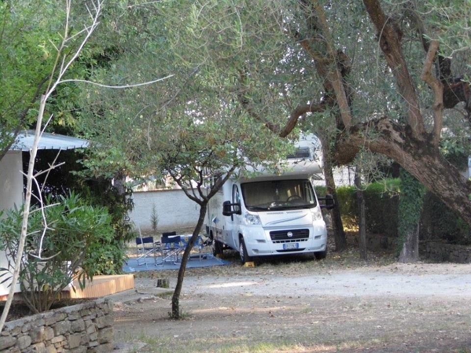 Weißes Wohnmobil auf einem Stellplatz am Campingplatz, umgeben von Bäumen und Hecken, mit Kiesfläche und Steinmauer im Hintergrund.