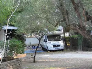 Weißes Wohnmobil auf einem Stellplatz am Campingplatz, umgeben von Bäumen und Hecken, mit Kiesfläche und Steinmauer im Hintergrund.