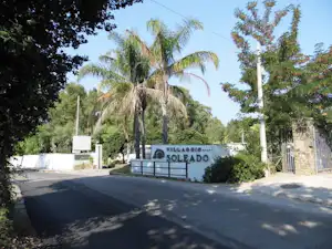 Einfahrt zum Campingplatz mit Schild „Villaggio Soleado“, Asphaltweg, Palmen und Bäumen am Eingang.