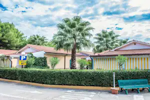 Camping-Bungalows mit gelb gestreifter Terrasse, Hecke und Palmen im Garten, daneben eine blaue Bank am Weg.