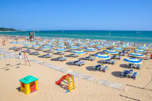 Strand mit vielen blauen und gelben Sonnenschirmen, Liegen und einem Spielbereich am Sand, Blick auf das Meer.
