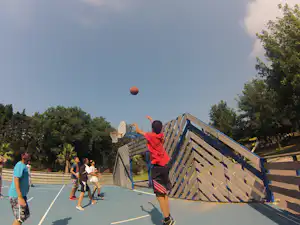 Basketballkorb auf dem Sportplatz eines Campingplatzes, Kinder spielen auf dem Asphalt zwischen Bäumen und Zaun.