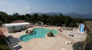 Schwimmbecken auf dem Campingplatz mit Sonnenliegen, gepflasterter Terrasse, Gewächshaus und Bäumen im Hintergrund.