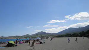 Zelt und Strand am See mit vielen Badegästen, im Hintergrund Berge und bewölkter Himmel.
