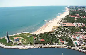 Luftaufnahme einer Küste mit Sandstrand, Campinganlage und Gebäuden am Meer, umgeben von Bäumen und Wegen.