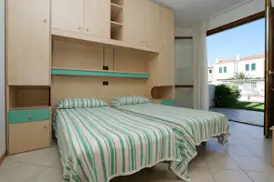 Schlafbereich mit Doppelbett, grün-weiß gestreifter Bettwäsche und Einbauschrank; Blick zur Terrasse mit Hecke und Fliesenboden.